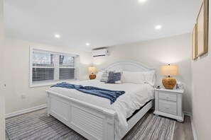 Un dormitorio con una cama blanca, dos mesitas de noche con lámparas, una ventana con persianas y un equipo de aire acondicionado en la pared.