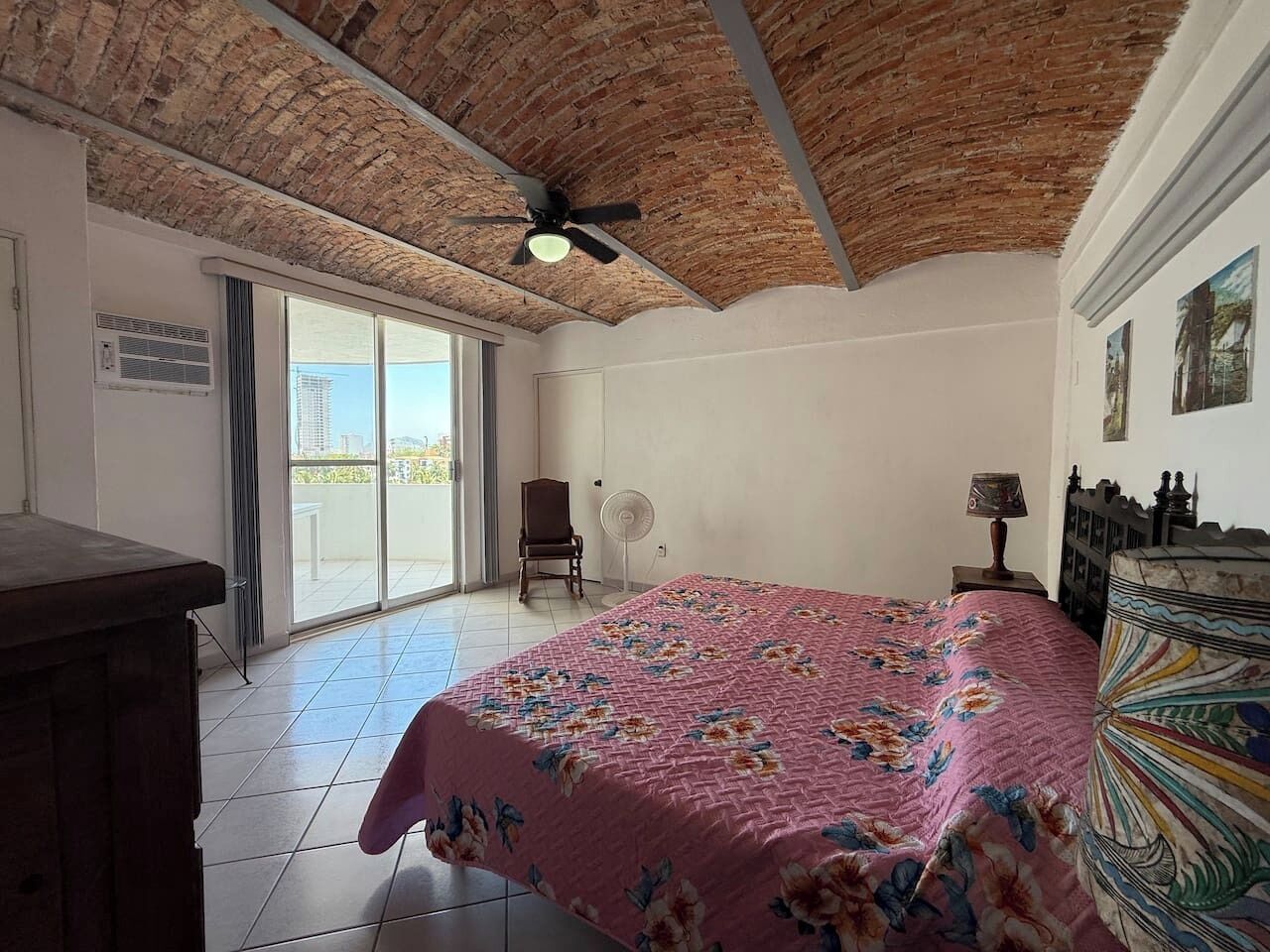 A bedroom with a brick ceiling, a bed with a floral bedspread, a chair, a fan, and a view through a glass door.