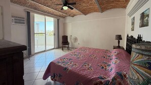 Ein Schlafzimmer mit Ziegeldecke, einem Bett mit floralem Überwurf, einem Sessel, einem Ventilator und Blick durch eine Glastür.