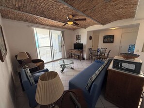 Ein Wohnzimmer mit einem blauen Sofa, einem Glastisch, einem Deckenventilator und einem Fernseher auf einem Holzboden.