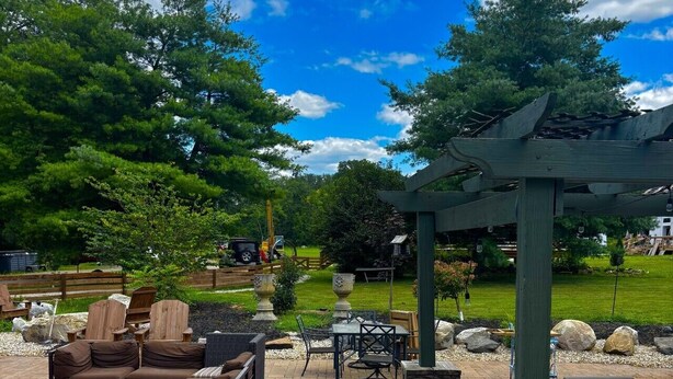 An outdoor patio with wicker furniture, a pergola, and a view of a grassy area with trees. - The Ruby Ranch House nestled behind Peyton Lake (Indianapolis)