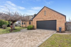A brick building with a garage door, a paved driveway, and a small garden area with a tree and shrubs. - Meadowstone Vista - Wanaka Holiday Home (Wanaka)