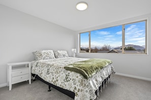 Quarto com cama, mesa de cabeceira e uma grande janela com vista para as montanhas.