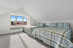 A bedroom with a sloped ceiling, two beds with plaid bedding, a large window with a view of mountains, and a built-in bench under the window. - Meadowstone Vista - Wanaka Holiday Home (Wanaka)