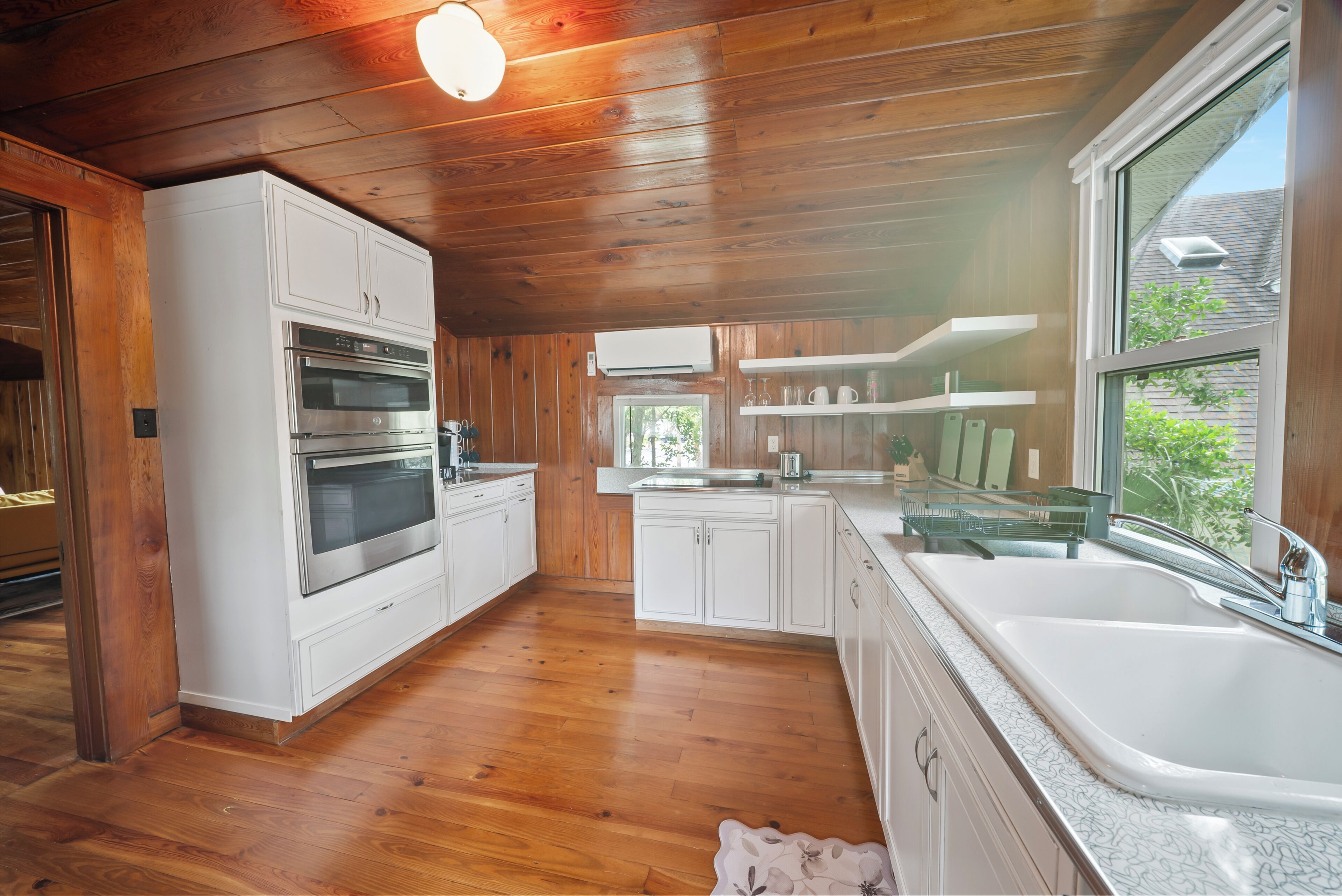 Een keuken met houten kasten, een dubbele oven, een spoelbak en een raam met uitzicht op groen.