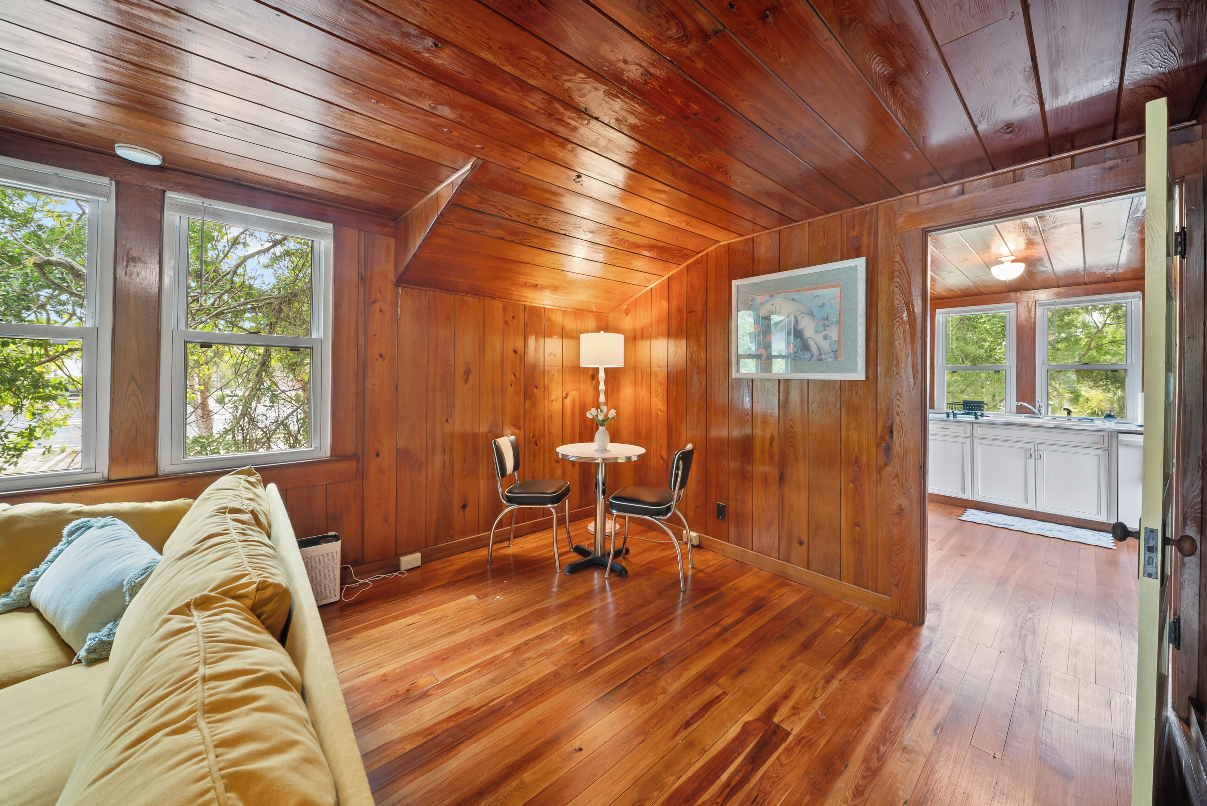 A cozy room with wooden walls, a wooden ceiling, a sofa, two chairs, a table with a lamp, and a window with a view of greenery.