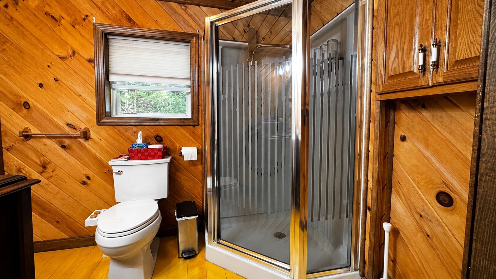 A wooden-paneled bathroom with a toilet, a shower enclosure, and a window.