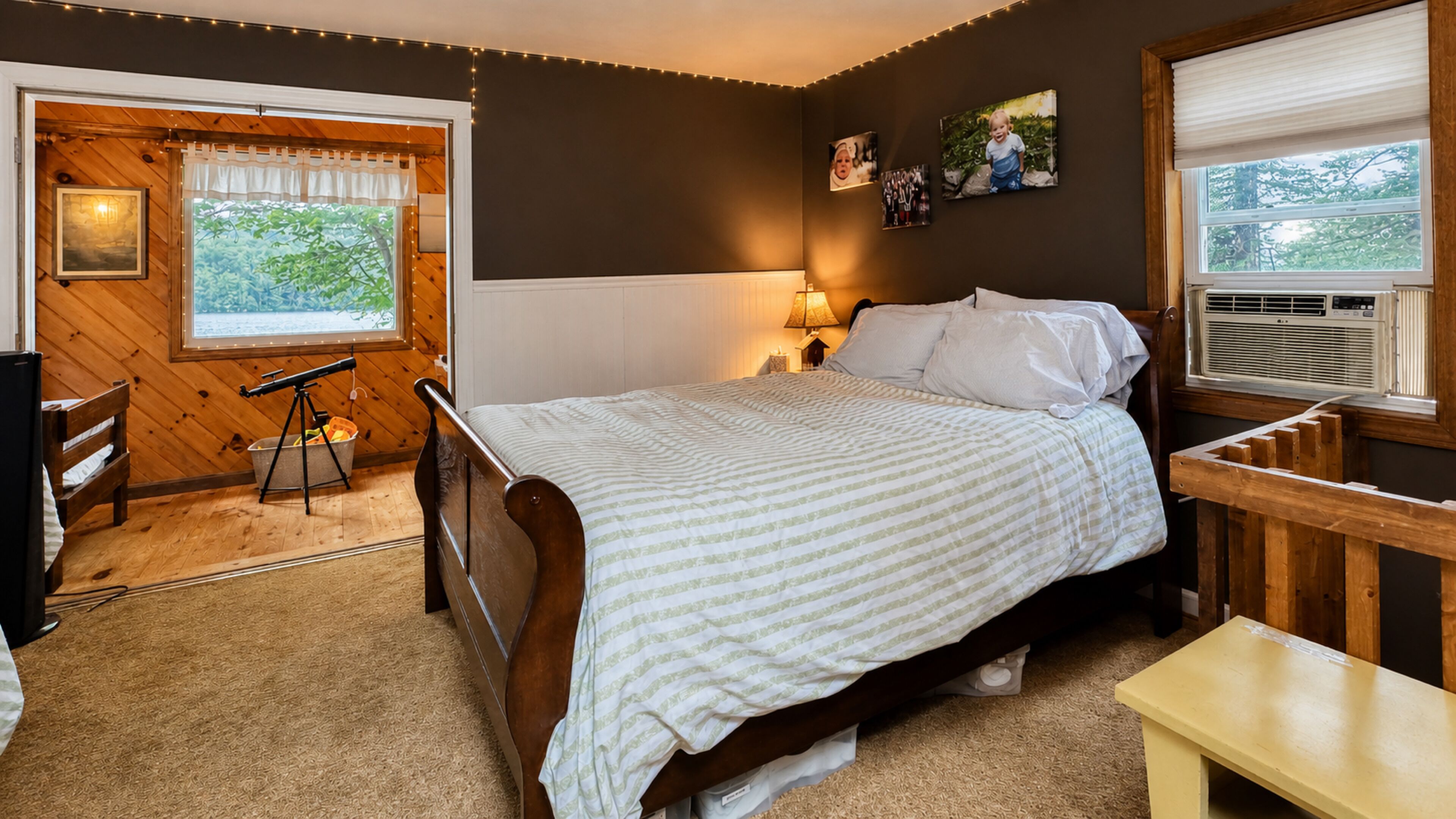 Un dormitorio con cama de madera, un escritorio, una silla, un ventanal con cortinas, una lámpara de pared y un marco de foto.
