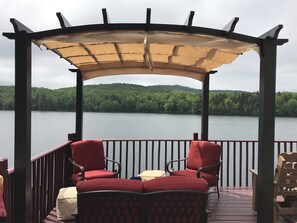 A covered outdoor seating area with two red chairs and a table, overlooking a lake with a forested hill in the background.