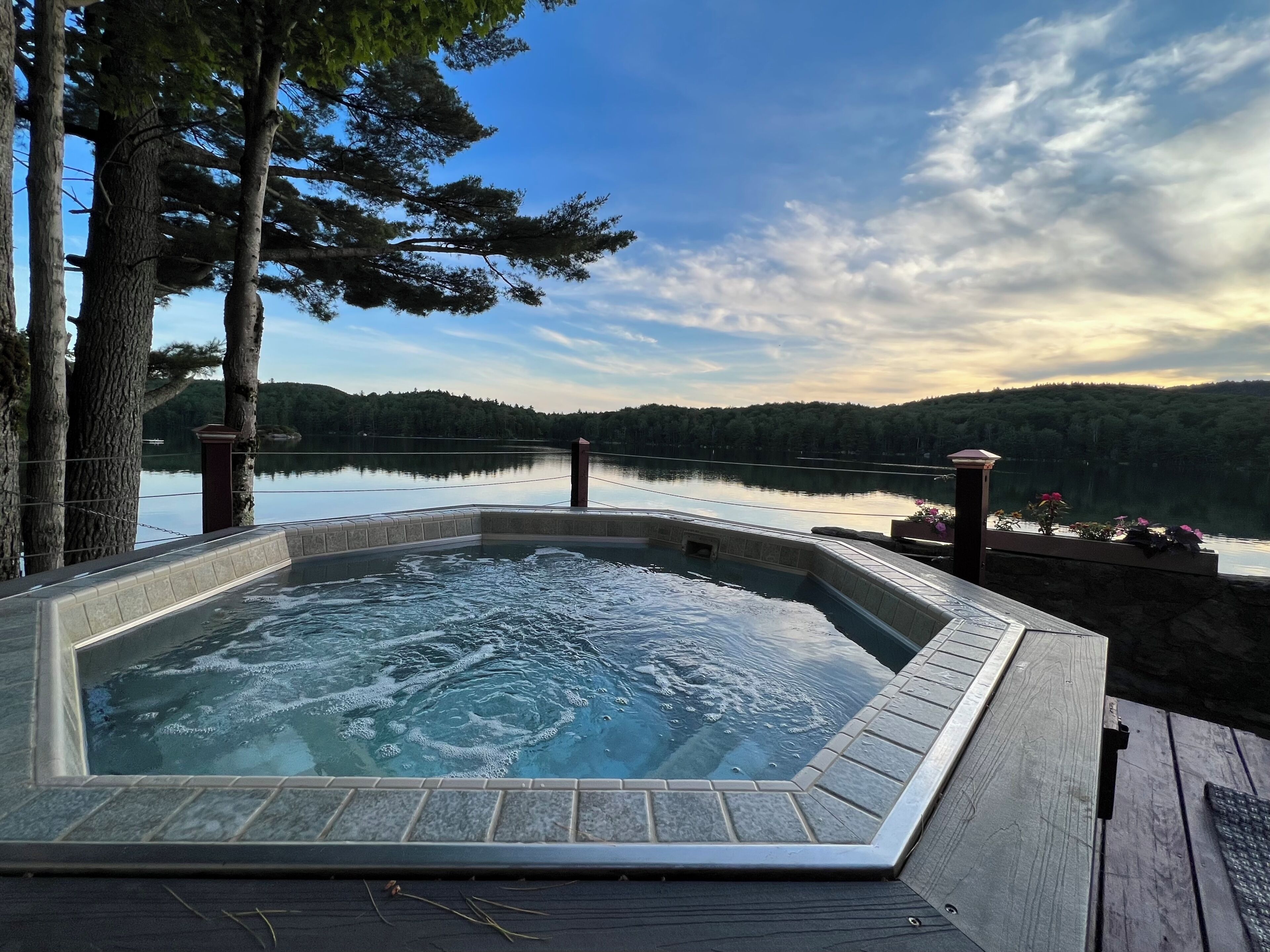 Un jacuzzi rodeado por una terraza de madera, con vistas a un lago tranquilo y colinas con árboles al fondo.