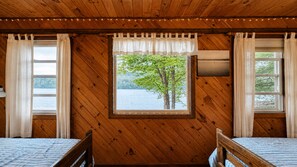 A cabin room with a wooden interior, two beds, a window with a view of a tree and water, and curtains.