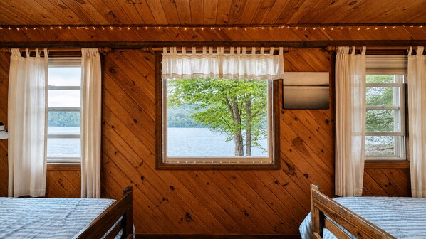A cabin room with a wooden interior, two beds, a window with a view of a tree and water, and curtains.