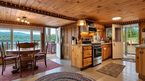 A wooden cabin interior with a dining area, kitchen, and large windows overlooking a forested landscape.