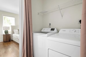 A laundry room with a front-loading washer and dryer, a drying rack, and a window with blinds. - Brand New - Downtown 2 Bedroom (Charleston)