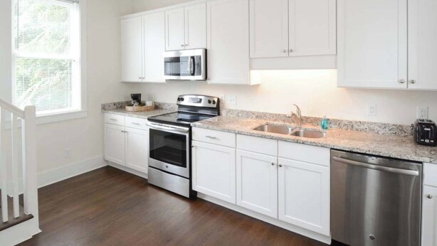 A kitchen with white cabinets, stainless steel appliances, a granite countertop, and a window with blinds. - Brand New - Downtown 2 Bedroom (Charleston)