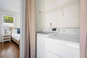 Une buanderie équipée d’un lave-linge et d’un sèche-linge à chargement frontal, d’une fenêtre avec des stores et d’un lit doté d’une tête de lit blanche et d’oreillers bleus.