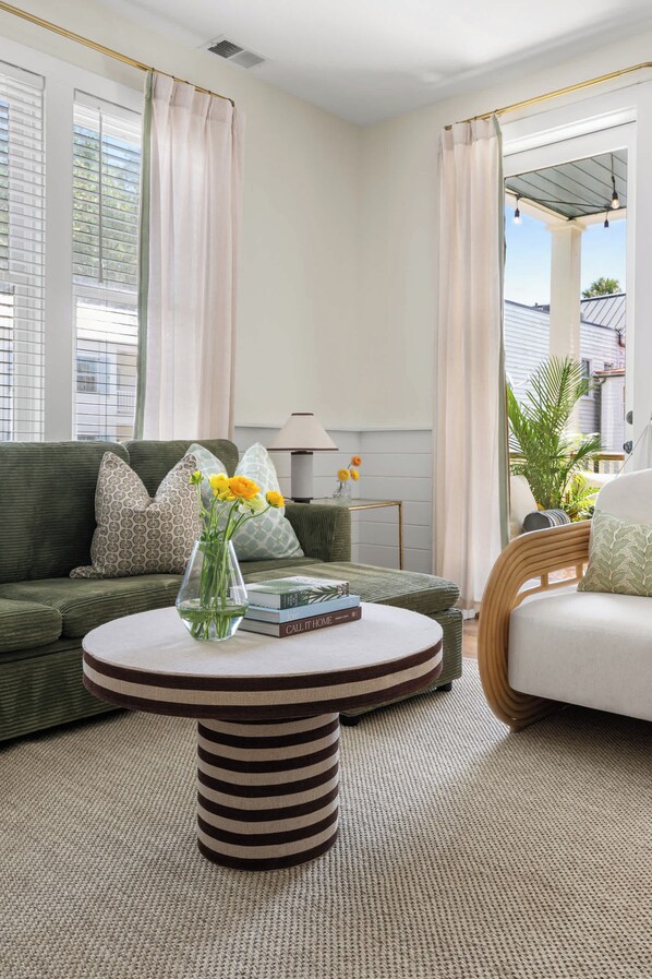 Una sala de estar con un sofá verde, una mesa de centro redonda y un sillón blanco. Hay una ventana con cortinas y vistas a edificios.