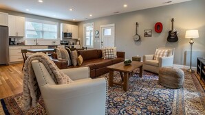 Uma sala de estar moderna com sofá, poltrona e mesa de centro, uma cozinha com armários brancos e uma área de refeições.