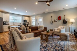 Ein modernes Wohnzimmer mit Sofa, Sessel und Couchtisch, eine Küche mit weißen Schränken sowie ein Essbereich.