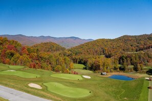 A golf course with a pond, surrounded by autumn-colored trees and mountains in the background. - Balsam Mountain Tree House | Luxury Retreat | Golf & Exclusive Club Amenities (Sylva)