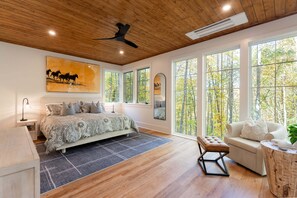 A bedroom with a large bed, multiple windows, a ceiling fan, and a view of trees. - Balsam Mountain Tree House | Luxury Retreat | Golf & Exclusive Club Amenities (Sylva)