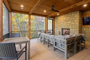 A wooden deck with a stone fireplace, seating area, and a television mounted on the wall. - Balsam Mountain Tree House | Luxury Retreat | Golf & Exclusive Club Amenities (Sylva)