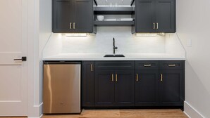 A modern kitchen with dark cabinetry, a stainless steel refrigerator, and under-cabinet lighting. - Balsam Mountain Tree House | Luxury Retreat | Golf & Exclusive Club Amenities (Sylva)