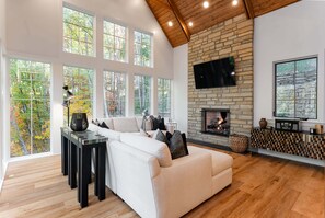 A spacious living room with a fireplace, large windows, and a high ceiling. - Balsam Mountain Tree House | Luxury Retreat | Golf & Exclusive Club Amenities (Sylva)