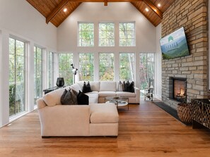 A spacious living room with a large sectional sofa, a fireplace, and large windows offering a view of the outdoors. - Balsam Mountain Tree House | Luxury Retreat | Golf & Exclusive Club Amenities (Sylva)