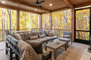 A covered outdoor seating area with a sofa, chairs, and a table, overlooking a forest with autumn foliage. - Balsam Mountain Tree House | Luxury Retreat | Golf & Exclusive Club Amenities (Sylva)
