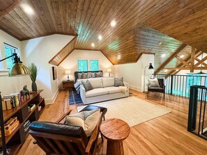 A cozy loft bedroom with a sloped ceiling, a bed, a sofa, a dining table, and a bookshelf. - Balsam Mountain Tree House | Luxury Retreat | Golf & Exclusive Club Amenities (Sylva)