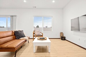 A modern living room with a leather sofa, a white coffee table, and a chair. There are two windows with blinds, a large TV on the wall, and a wooden floor. - Cheney Home | Close to Interstate & Airport (Cheney)
