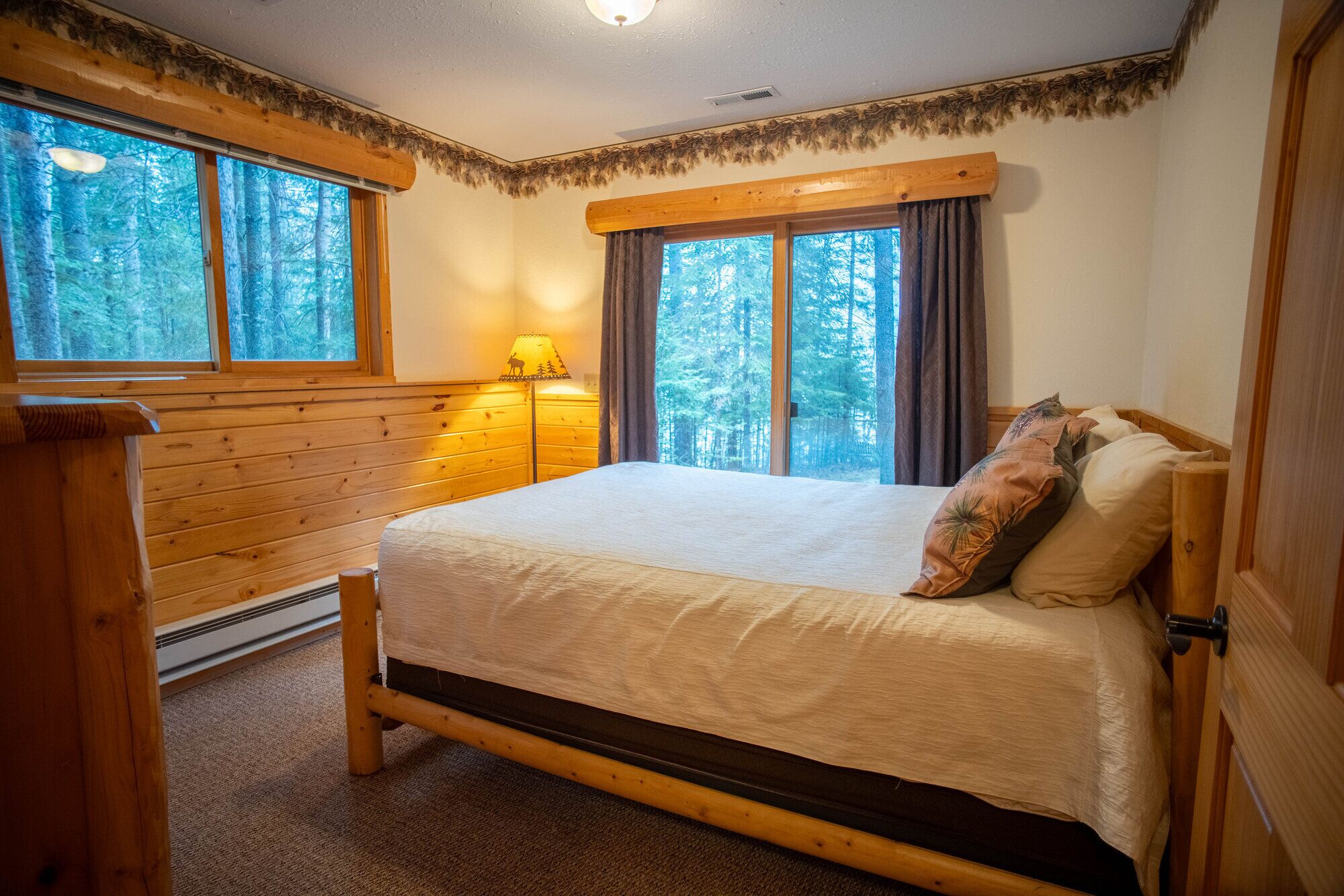 Un lit en bois avec un matelas blanc et des coussins décoratifs, une table de chevet, une lampe et une vue sur une forêt à travers de grandes fenêtres.
