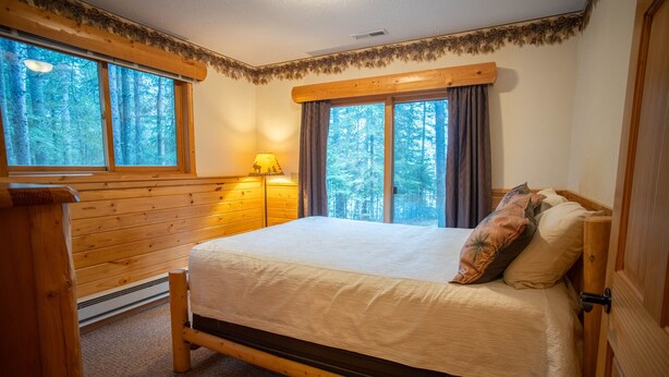 A wooden-framed bed with a white mattress and decorative pillows, a nightstand, a lamp, and a view of a forest through large windows. - The "Grand Dame" of Timber Bay (Babbitt)