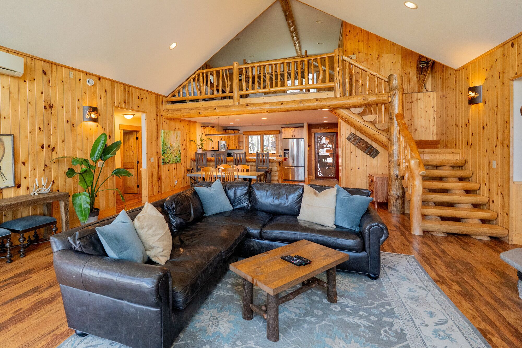 A log cabin interior with a leather sectional sofa, a wooden coffee table, a staircase, and a kitchen area.