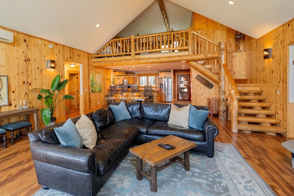 A log cabin interior with a leather sectional sofa, a wooden coffee table, a staircase, and a kitchen area.