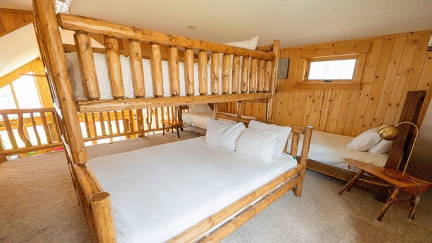 A wooden cabin room with a bunk bed, a single bed, a small table, and a window. - The exclusive "Owner’s Edition": The Hefty Logger Lodge (Babbitt)