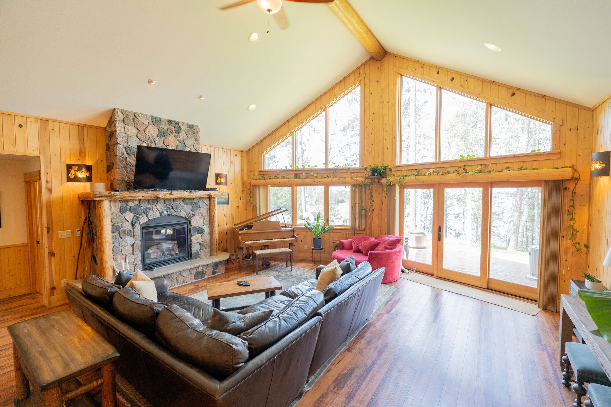 A spacious living room with a stone fireplace, a grand piano, a leather sofa, and large windows offering a view of the outdoors.