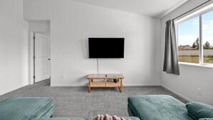 Una stanza con una TV a schermo piatto montata a parete, un tavolo in legno, due divani verdi e una finestra con vista sull'esterno.