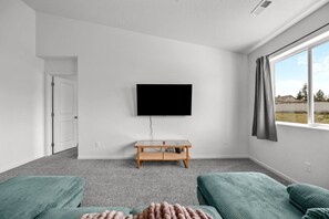 A room with a flat-screen TV mounted on the wall, a wooden table, two green sofas, and a window with a view of the outdoors.
