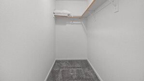 Una habitación estrecha con paredes blancas, piso alfombrado en gris, una estantería de madera con un cojín y un techo blanco con una viga de madera.