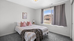 Una camera da letto con un letto, un comodino, una lampada, una finestra con tende e vista su una casa.