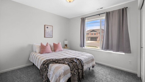 Un dormitorio con cama, mesita de noche, lámpara, ventana con cortinas y vista a una casa.