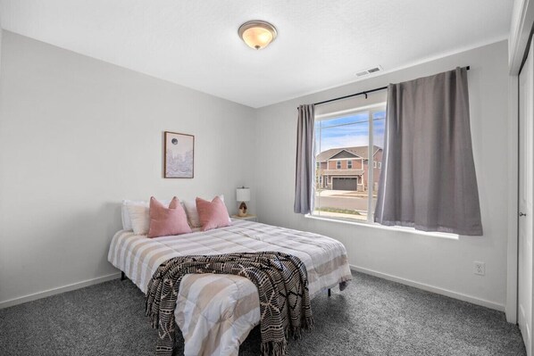 A bedroom with a bed, a bedside table, a lamp, a window with curtains, and a view of a house outside.