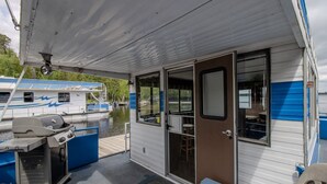 Een overdekt terras met een barbecue, een deur naar binnen en uitzicht op een rivier met boten.
