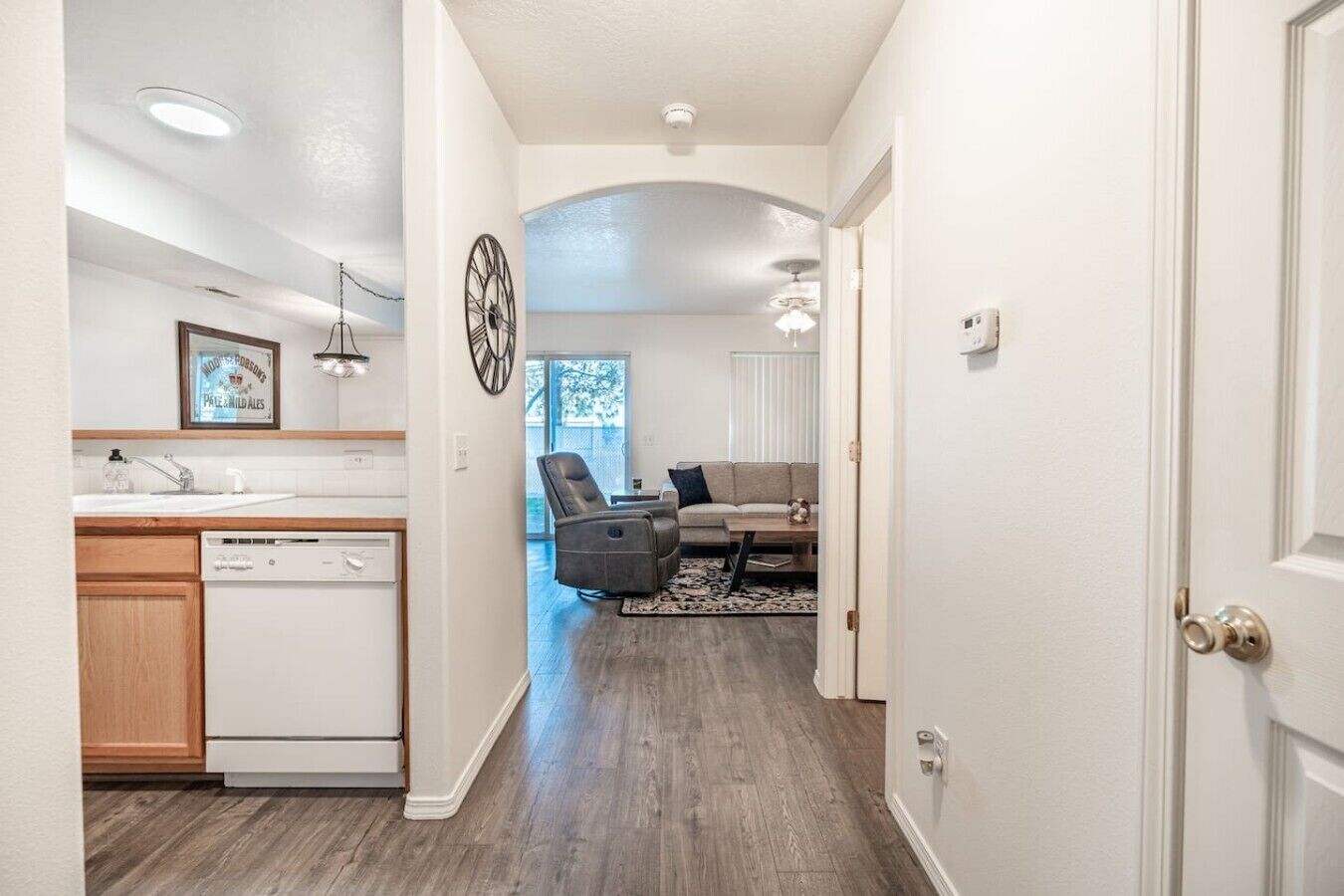 A modern kitchen with a dishwasher, a dining area with a sofa and a coffee table, and a hallway leading to a living room with a fireplace.