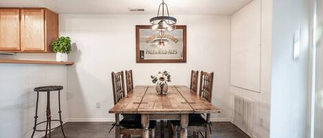 A dining area with a wooden table, chairs, a hanging light fixture, and a framed sign on the wall.