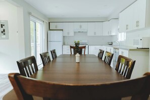 Uma sala de jantar com uma longa mesa de madeira rodeada por cadeiras, uma planta central e armários brancos ao fundo.