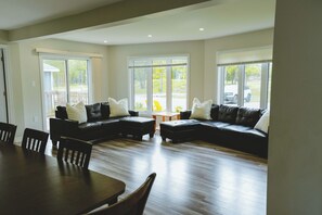 A spacious living room with a sectional sofa, a dining table, and chairs. There are large windows providing views of the outdoors. - Leo Cottage - 5-Bedroom Cottage – Family Friendly (Trent Lakes)