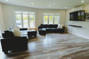 A modern living room with a sectional sofa, a flat-screen TV mounted on the wall, and large windows providing natural light. - Leo Cottage - 5-Bedroom Cottage – Family Friendly (Trent Lakes)